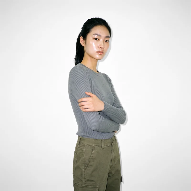 A 21-year-old Korean woman wears women's olive utility cargo pants in a three-quarter-length frame, shown from a front three-quarter angle in a self-hug pose with a soft facial expression. A Seasalt studio-wall backdrop fills the background.