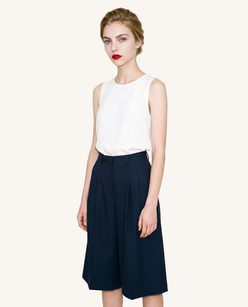 An 18-year-old Caucasian female model stands straight in an angled front view, shown from three-quarter length, wearing navy pleated culottes. The garment is presented against a soft ivory flat-color backdrop.