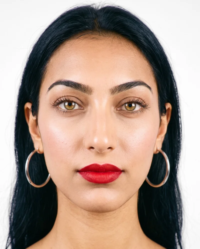 A 19-year-old Arab woman is shown from the head in a front view with her head level and a neutral expression, wearing silver hoop earrings. A cloud-white studio-wall backdrop frames the composition.