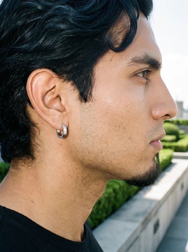 A side view tightly frames the back of a male ear wearing a men's huggie hoop earring. The ear is shown against a rooftop garden formal backdrop.