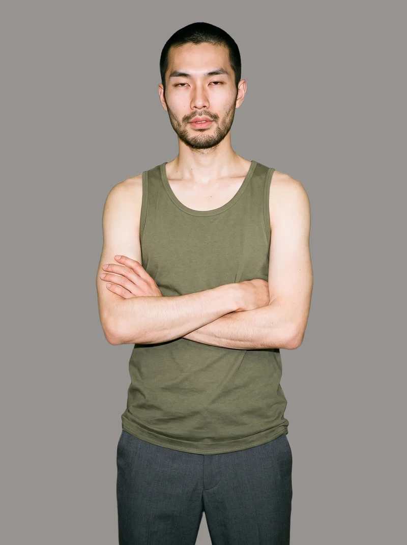 A 32-year-old Japanese man wears an olive cotton tank top in a three-quarter-length front view. He poses with a self-hug and a soft facial expression against a flat grey studio backdrop.