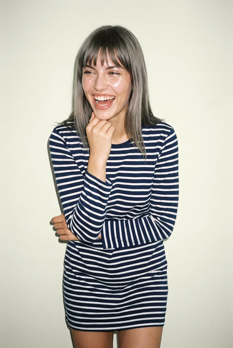 A 20-year-old Slavic woman models a striped nightdress in an upper-body front view. She is laughing with one wrist raised at her jaw, against an ivory lace backdrop.