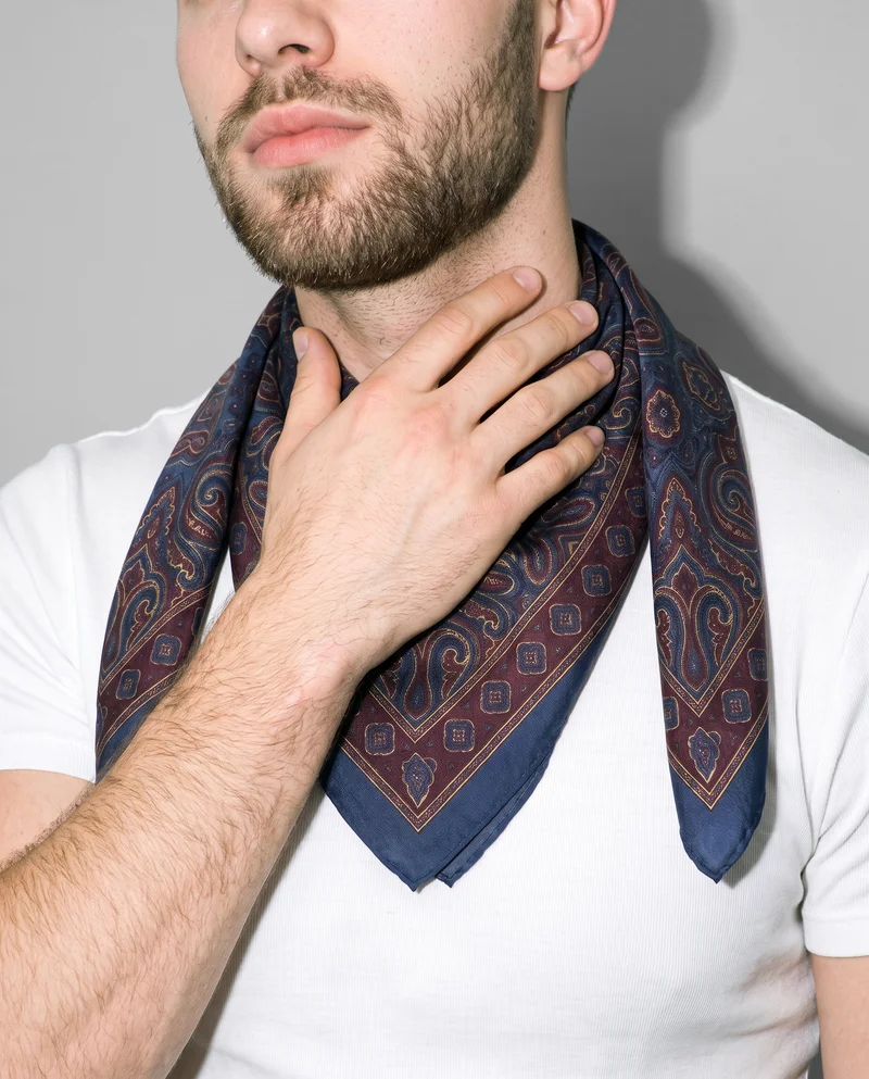 A male model wears a patterned silk scarf around his neck, shown from the front with one hand resting on his neck. The image is framed closely around the neck and scarf against a dove grey studio backdrop.