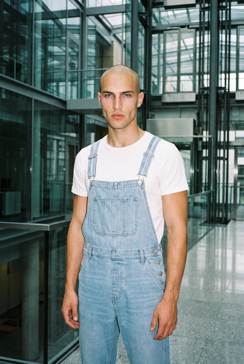 A 20-year-old Caucasian male model wears light wash denim dungarees in a relaxed standing pose. He is shown from the front in a three-quarter-length frame with a neutral expression, against a skyscraper glass atrium backdrop.