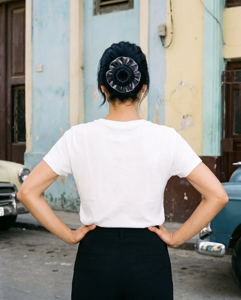 A 22-year-old Turkish woman is shown from the rear in an upper-body view with her hands on her hips, wearing a velvet hair tie. The backdrop features vintage cars on a Cuban street.