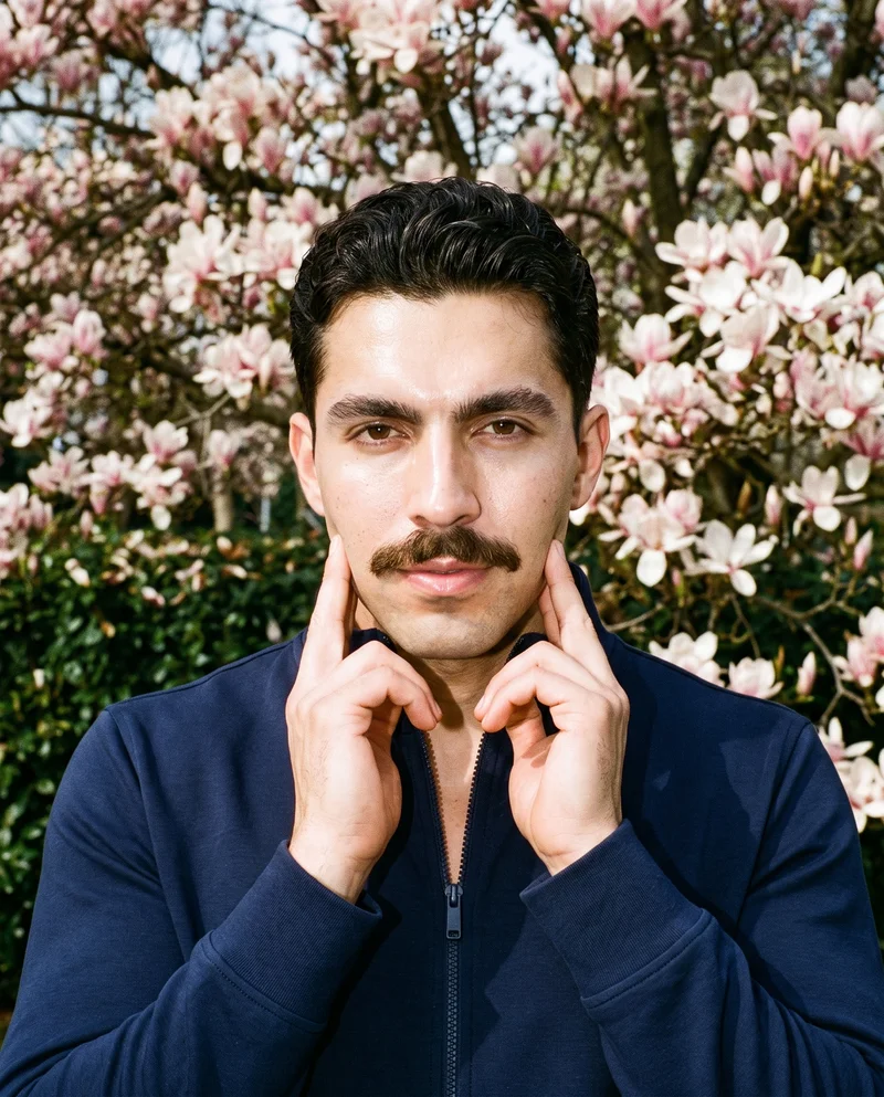 A 24-year-old Hispanic man is shown in a bust-length front view wearing a navy tracksuit. His hands are positioned at his jaw, with a confident expression, against a Magnolia Tree Park backdrop.