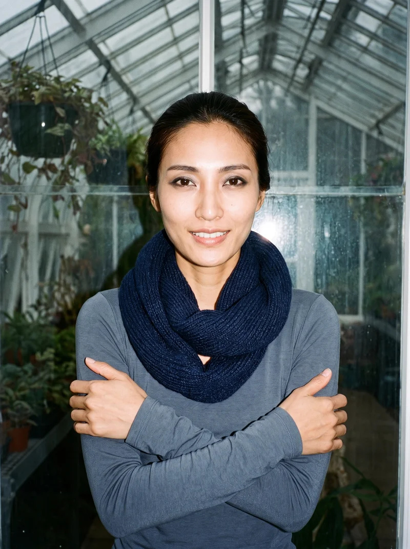 A 32-year-old Japanese woman is shown from the front in an upper-body view, smiling and holding herself in a self-hug pose while wearing a navy snood. A greenhouse conservatory serves as the backdrop.