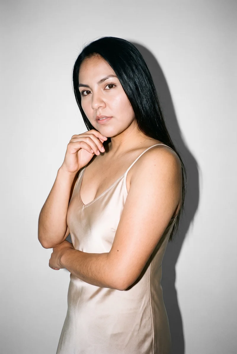 A 26-year-old Indigenous American woman wears a champagne slip dress in an upper-body frame, shown from a front three-quarter angle with one hand at her chin and a soft facial expression. A pearl-grey studio-wall backdrop fills the background.