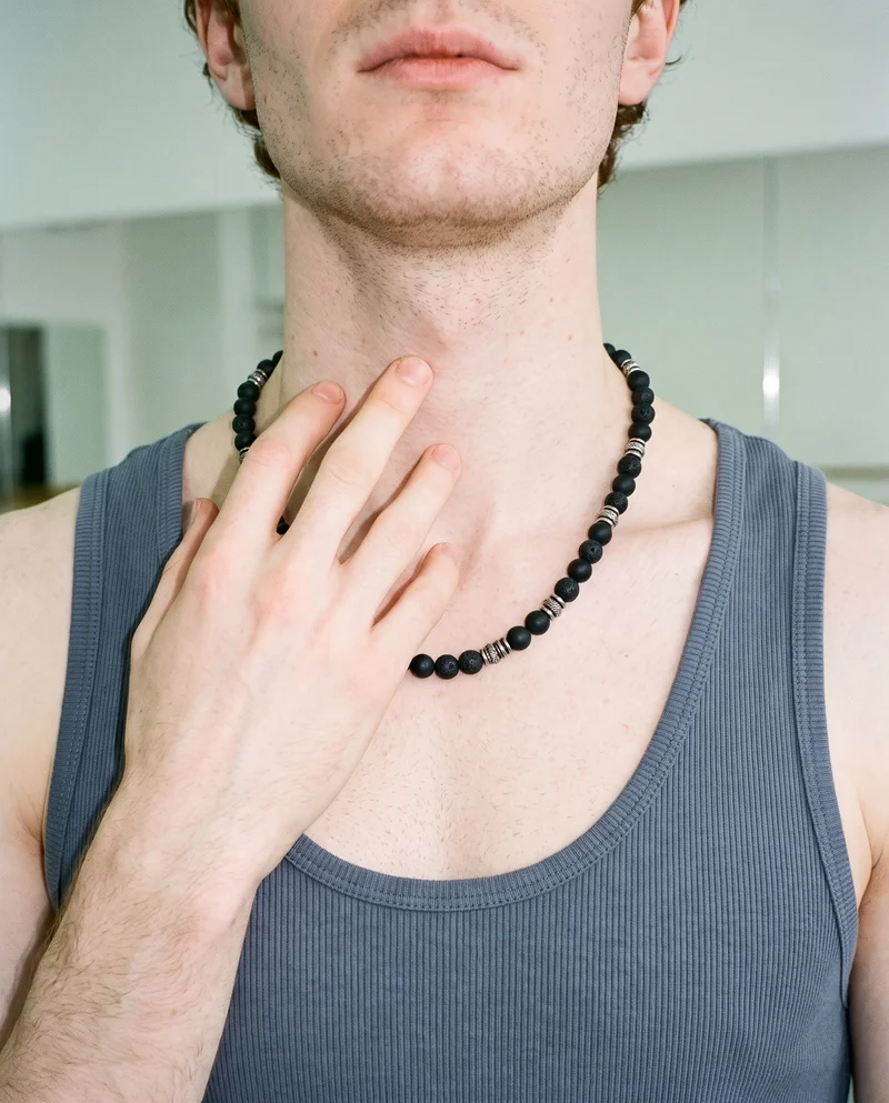 A man's neck is shown from the front with a beaded chain worn around it. His fingers rest down the front of his throat, with a dance studio backdrop behind him.