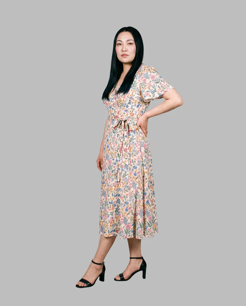 A 38-year-old Chinese woman wears a floral print wrap dress in a full-body view from a front three-quarter angle. One foot is positioned forward, and she has a serious facial expression. A dove grey flat-color backdrop fills the background.