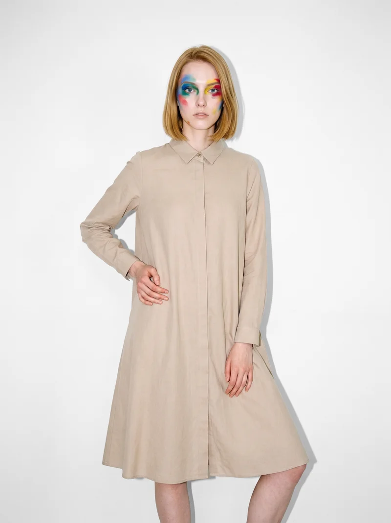 A 20-year-old Caucasian woman wears a beige modest shirt dress in a front-facing three-quarter-length view. She stands with her weight shifted back and a serious expression against a Seasalt studio-wall backdrop.