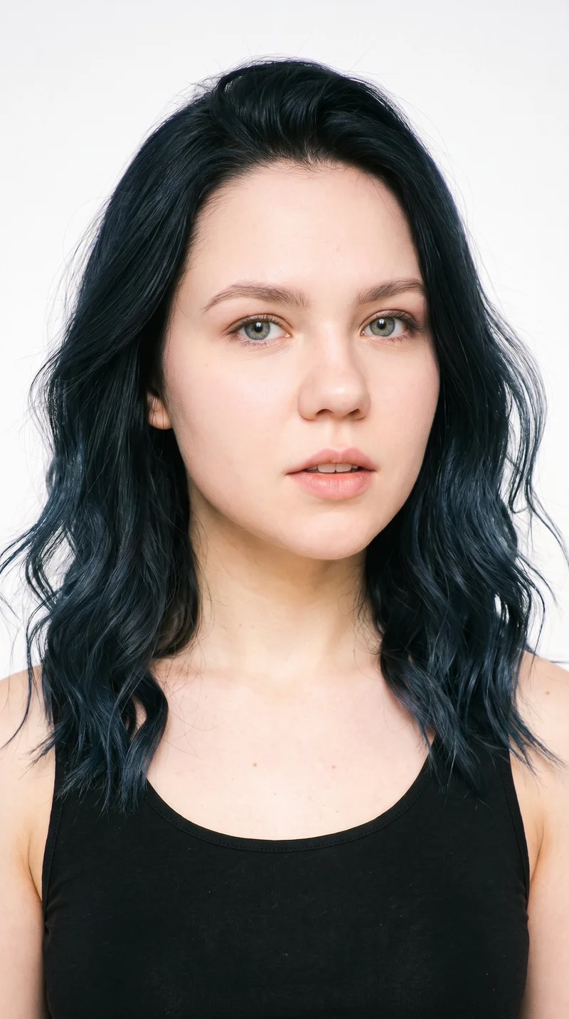 A 22-year-old Slavic woman with very light pale skin and a peach undertone has a youthful baby face with full cheeks and softly rounded contours. Her eyebrows are thin, and her eyes are blue with a cool undertone. She has thin lips and black hair styled in loose, slightly tousled beach waves with natural volume. No facial hair is recorded.