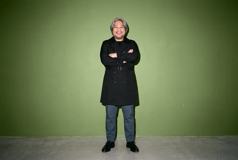 A 45-year-old Southeast Asian man wears a black trench coat in an environmental front-view composition. He stands with his arms crossed and a smile, against an olive grove backdrop.