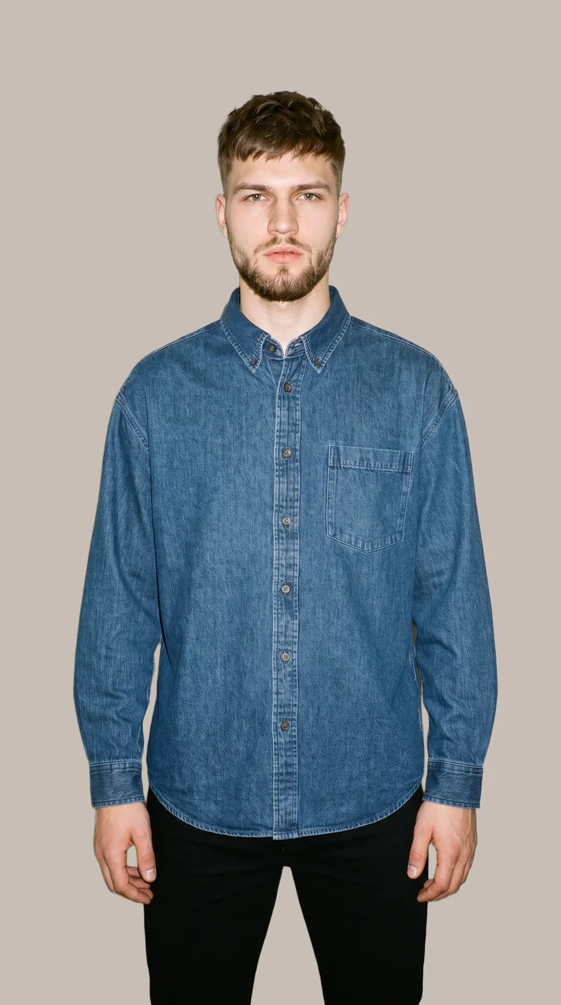 A 22-year-old Slavic man wears an oversized denim shirt in an upper-body, front-view composition. His arms are relaxed and his expression is neutral, set against a flat greige backdrop.