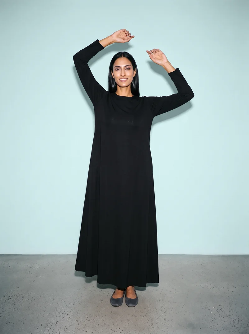A 34-year-old Arab woman models a black classic abaya in a full-body, front-facing view. She is smiling with her arms overhead, against a duck-egg studio wall backdrop.