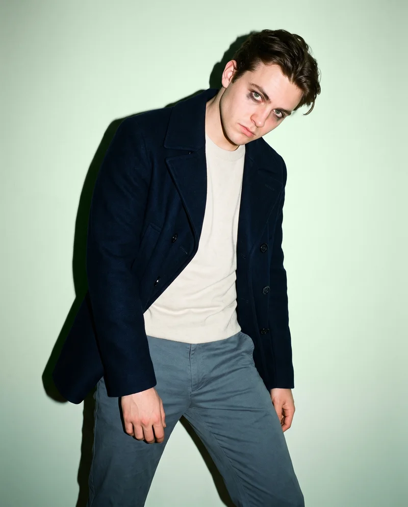 A 20-year-old Caucasian man wears a navy double-breasted wool coat in a three-quarter-length front view. He poses with one hip out and an intense expression against a pistachio studio-wall backdrop.