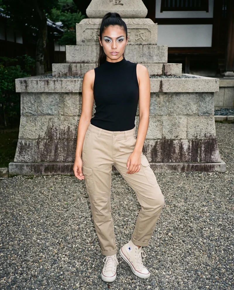 A 20-year-old Hispanic woman models women's khaki hiking trousers in a full-body front view, standing with her weight back and an intense expression. A Kyoto temple garden forms the backdrop.