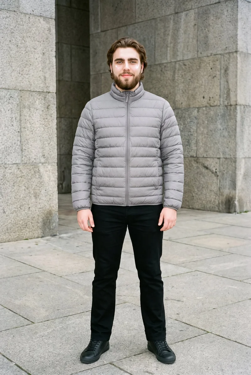 A 19-year-old Slavic man is shown full body in a front view, standing straight with a natural expression and wearing a grey lightweight down jacket. A brutalist monument plaza forms the backdrop.