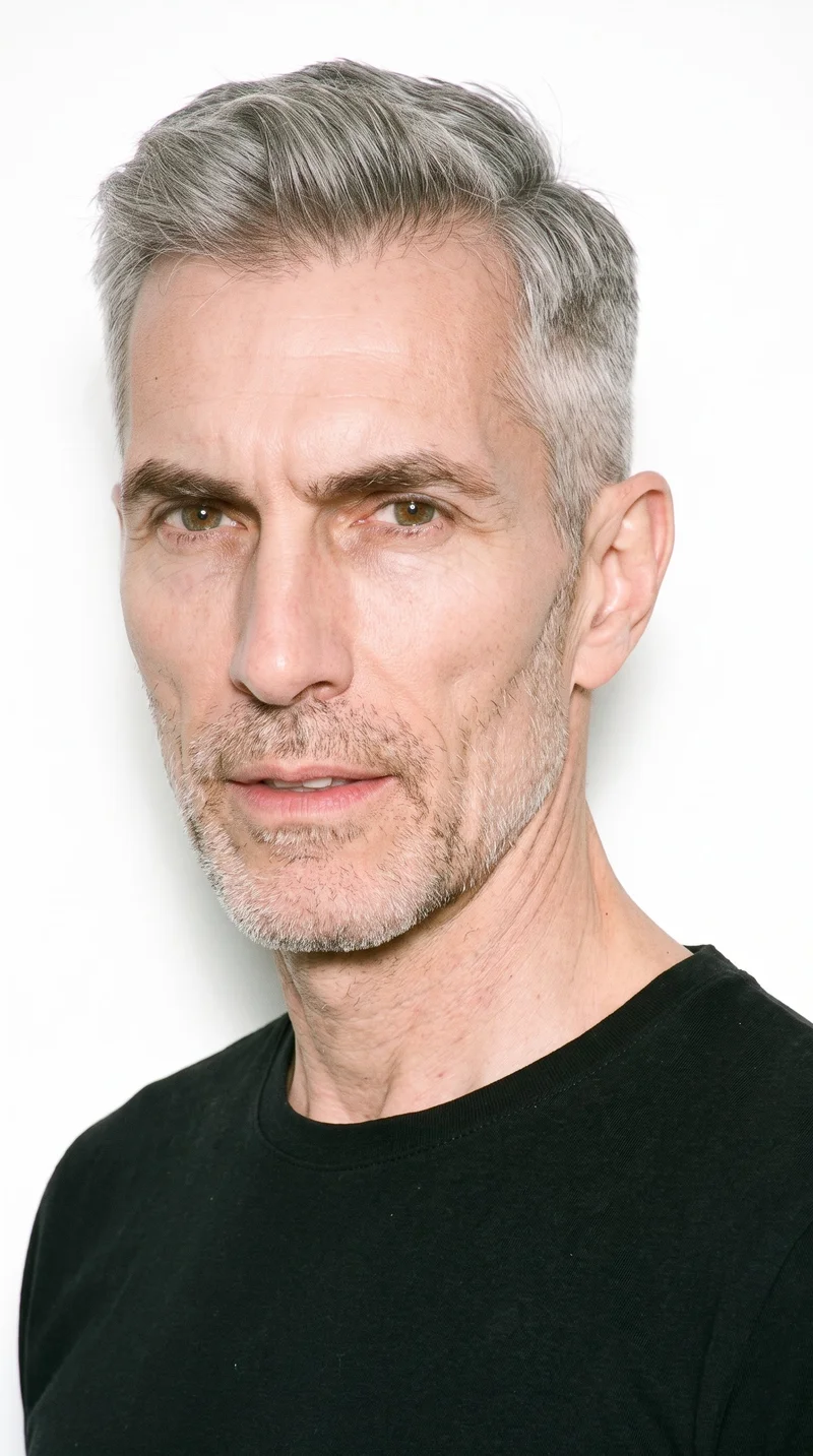 A 56-year-old Caucasian man with fair skin and a cool undertone has a gaunt face defined by lean features, hollowed cheeks, and sharply sculpted bone structure. His thick eyebrows frame hazel eyes with green-gold flecks, and his medium lips sit above dense, heavy stubble forming a dark, defined layer across the beard area. His gray hair is styled in a two-block cut, with short sides and back contrasted by a longer, fuller top.