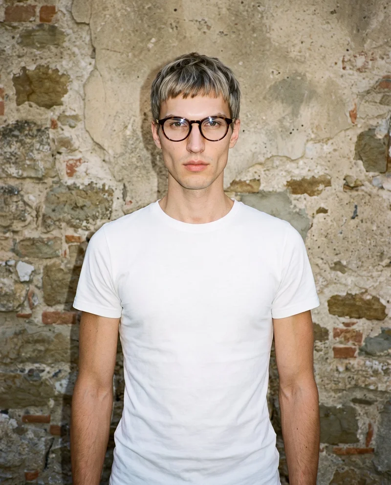 A 27-year-old Slavic man is shown from the upper body in a front-facing view, wearing round blue-light glasses. His arms are relaxed and his facial expression is neutral, with a Tuscany hilltop village backdrop.