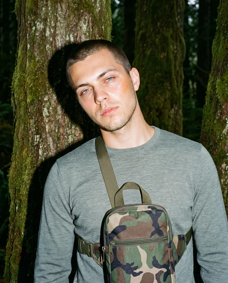 A 21-year-old Caucasian man is shown from the front in an upper-body view wearing a men's camo bum bag. He has a shoulder-drop pose and a soft facial expression, with an Olympic Rainforest backdrop.