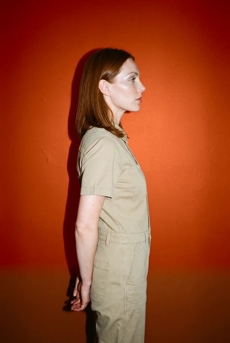 A 28-year-old Caucasian woman wears a khaki utility jumpsuit in an upper-body side-view composition. She is smiling with her hands behind her back, against a rust studio-wall backdrop.