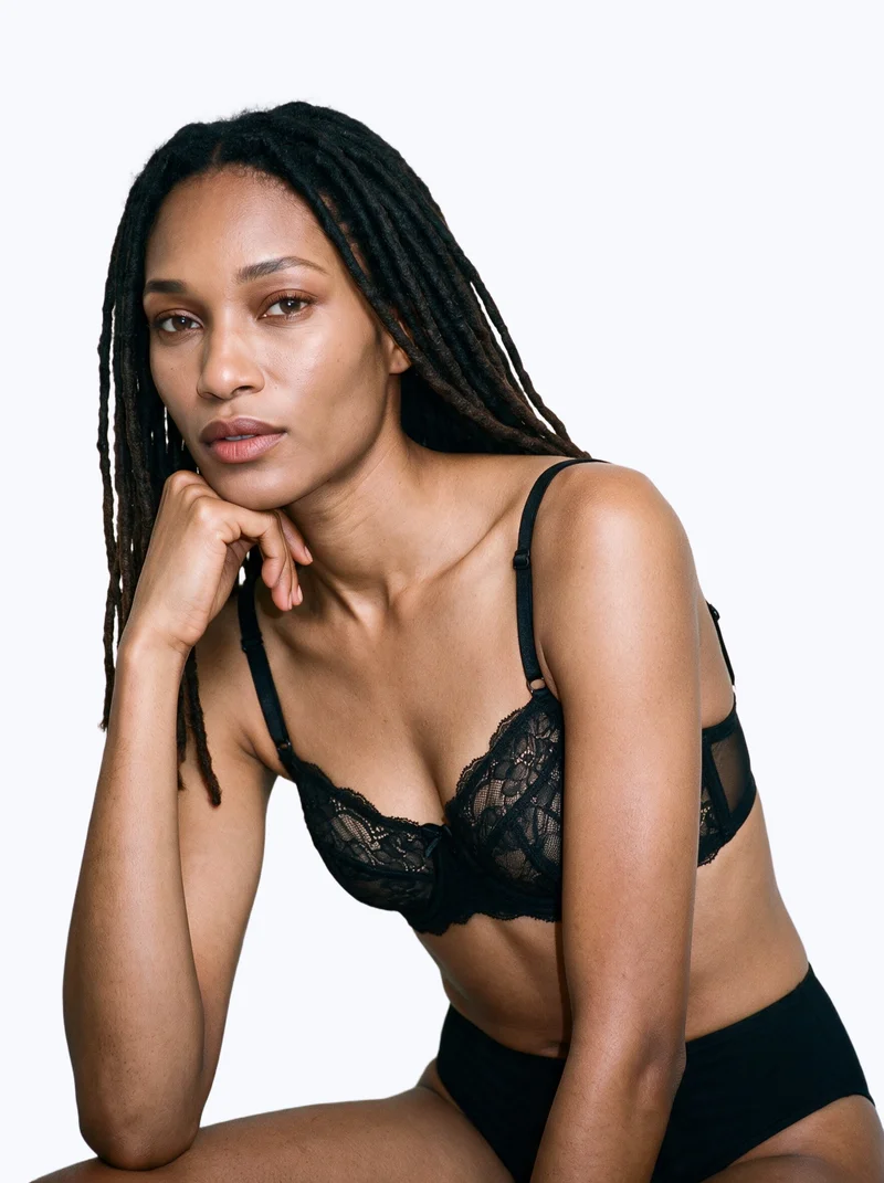 A 26-year-old African woman wears a black lace bra in an upper-body composition viewed from an angled front perspective. Her wrist is positioned at her jaw, and she has a confident facial expression against a flat porcelain-colored backdrop.