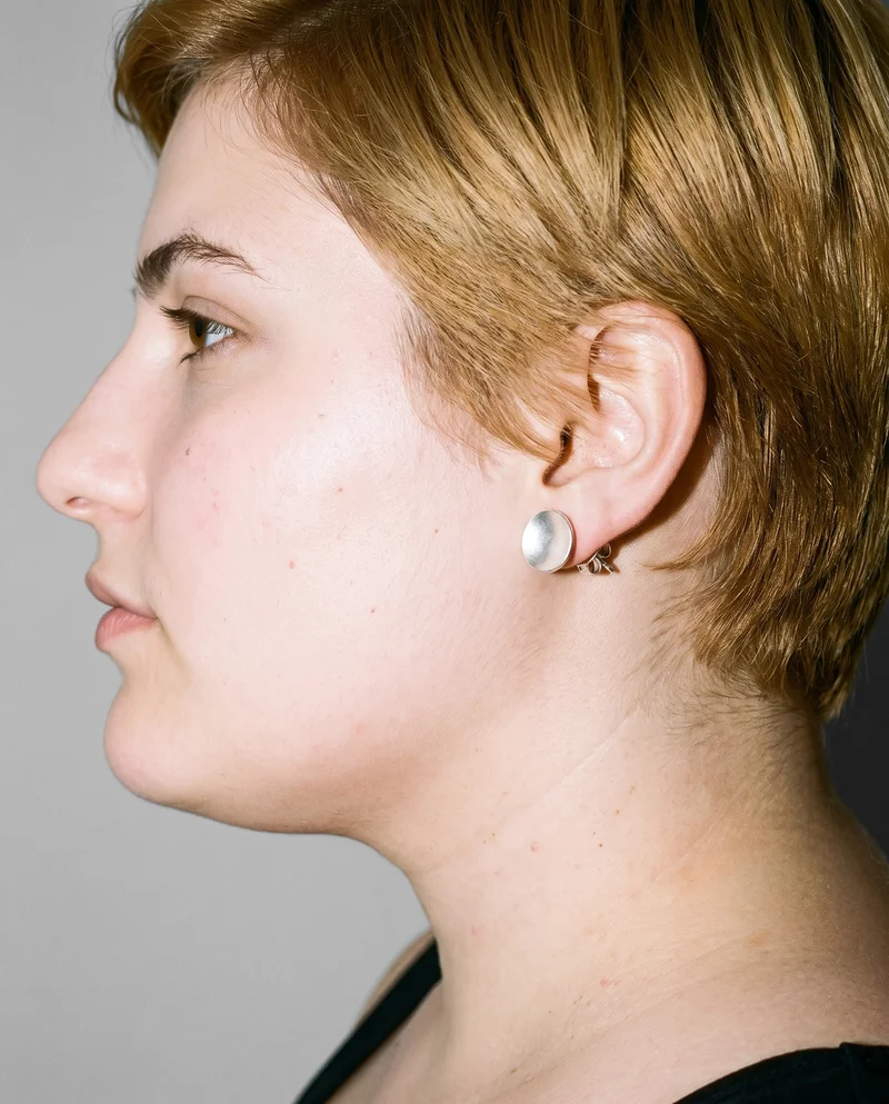 A woman's ear shown at ear level in side view, wearing a silver stud earring. A dove grey studio-wall backdrop fills the background.