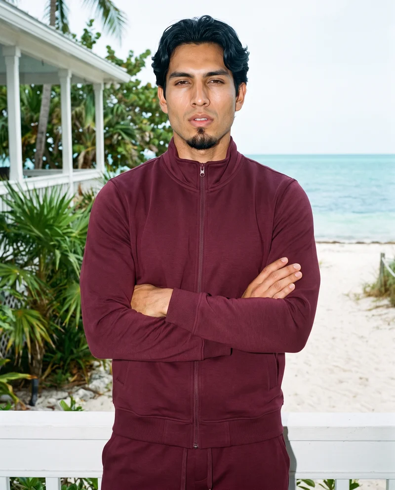 A 25-year-old Hispanic man wears a burgundy tracksuit in an upper-body front view, standing with his arms crossed and a soft facial expression. A Key West porch serves as the backdrop.