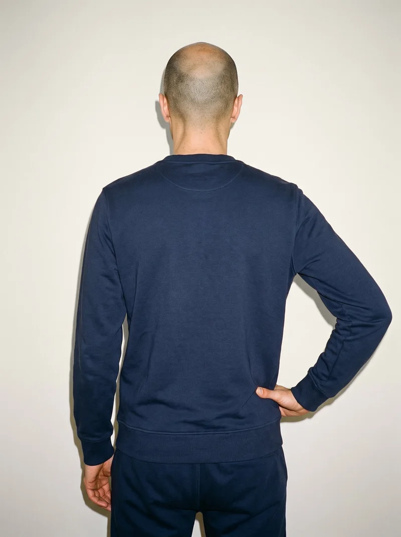 A 37-year-old Slavic man is shown from behind in an upper-body frame wearing a navy loungewear set, with one hand resting on his hip. An Oyster Pearl studio-wall backdrop is visible behind him.