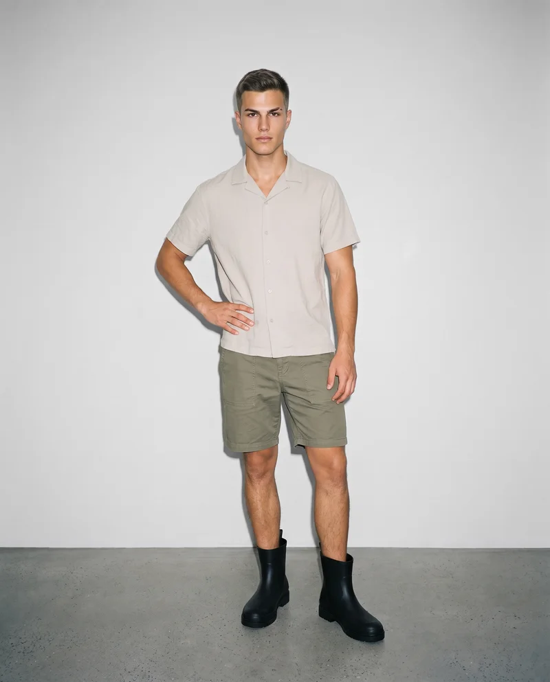 A 20-year-old Caucasian man is shown full-body from the front with one hand on his hip, wearing black rain boots. He has a confident facial expression, with a Fog Grey studio-wall backdrop.