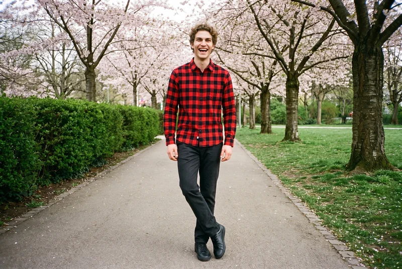 A 21-year-old Slavic man wears a red check flannel shirt in a front view, standing with his ankles crossed and laughing. Cherry blossoms form the park backdrop.