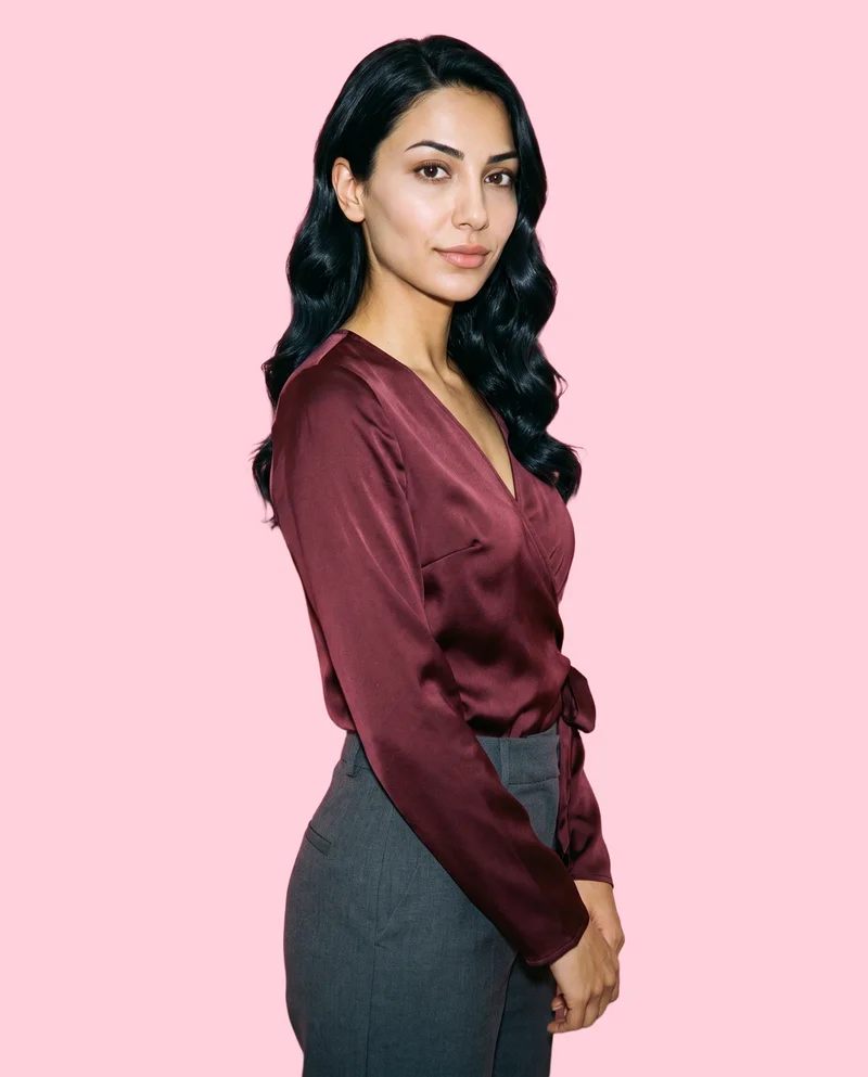 A 24-year-old Turkish woman wears a burgundy satin wrap top, shown from the upper body at a front three-quarter angle with her hands in front and a confident expression. The light powder pink backdrop is a flat color.