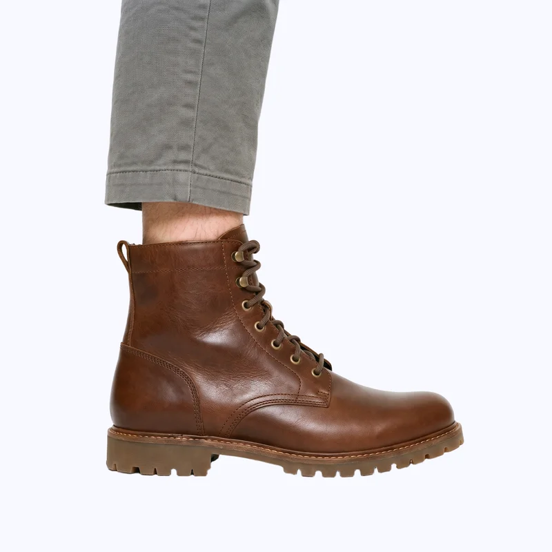Brown men's lace-up boots shown from the side at ankle level, framed around the ankle against a plain porcelain-colored backdrop.