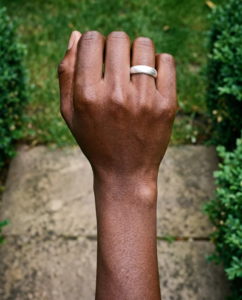 A closed hand and wrist shown from above, wearing a women's silver stacking ring. A topiary garden forms the backdrop.