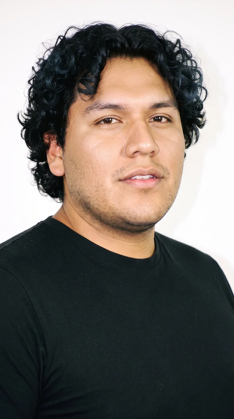 A 25-year-old Indigenous American man with medium olive skin and a greenish-golden undertone. His face has sharp, angular features, a defined jaw, and pronounced cheekbones. He has medium-thickness eyebrows, brown eyes, and full lips. His black hair forms defined, springy curls with natural volume, and fine, closely trimmed stubble lightly covers the beard area.