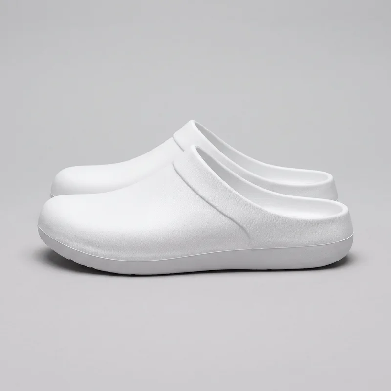 A matching pair of white closed-toe slip-on clogs is shown from a side three-quarter angle. The backless design has rounded enclosed toes, low-cut open heels, raised rims around the foot openings, molded side ridges, and slightly elevated platform-style soles with clean sidewalls.