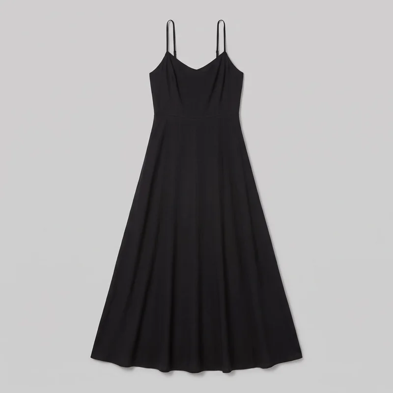A mostly complete black sleeveless camisole fit-and-flare maxi dress is laid flat against a plain backdrop. The dress has a V neckline, narrow adjustable spaghetti straps with visible sliders, curved arm openings, a tailored bodice with subtle vertical seams, a defined horizontal waist seam, and a wide A-line skirt ending in a softly wavy hem.