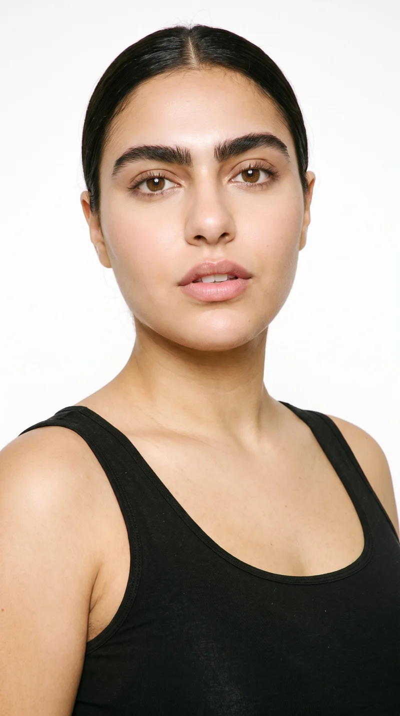 A 25-year-old Arab woman with a medium olive skin tone and greenish-golden undertone. Her round face has soft, full contours with gentle curves and few angles, framed by thick eyebrows, brown eyes, and medium-sized lips. Her dark-brown hair is smoothed back into a neat low bun at the nape.