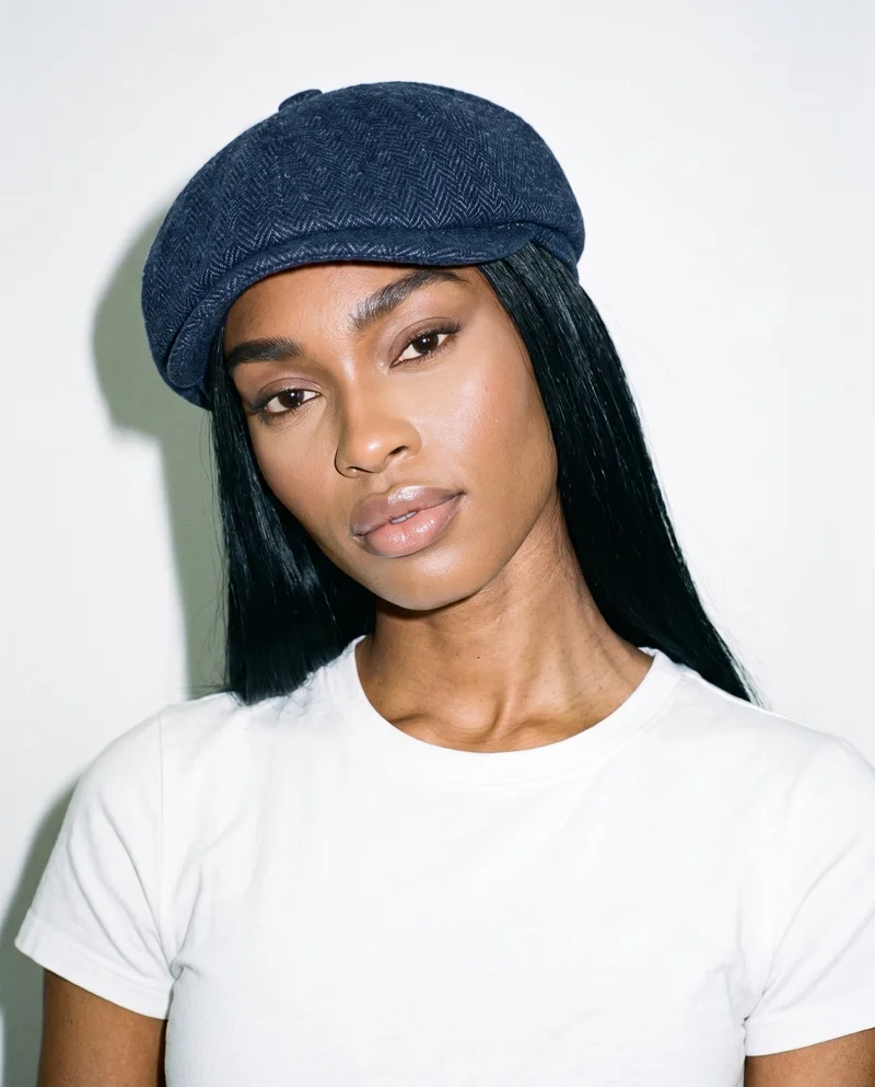 A 21-year-old African woman is shown from the bust up in a front view, wearing a women's navy herringbone flat cap. Her head is tilted with a confident expression. A museum gallery serves as the backdrop.