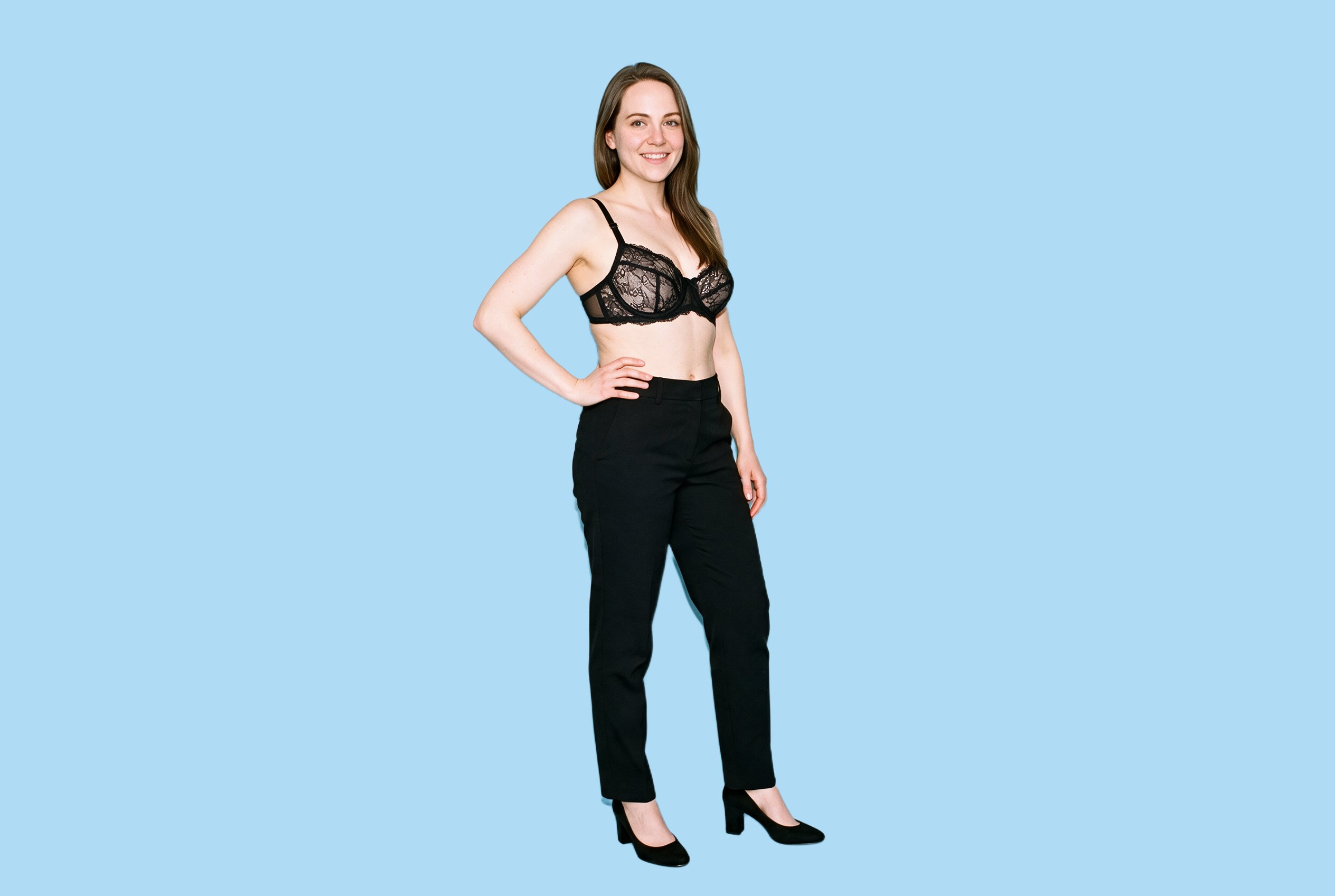 A 33-year-old Caucasian woman models a black lace bra in an angled front, full-body view. She stands with one hand on her hip and a natural facial expression against an ice-blue flat-color backdrop.