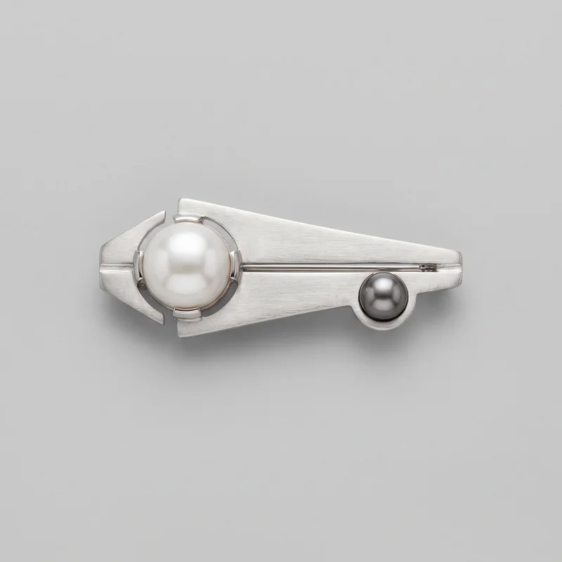 A geometric men's brooch is shown horizontally in a standalone close view. It has an elongated asymmetric bar silhouette with a tapered angular body, stepped pointed shaping at one end, and a rounded blunt end at the other. Fine linear brushing covers the surface, with a narrow horizontal pin-stem channel and a small clasp mechanism visible near one end. A large round pearl-like dome sits off-center in an open circular frame with four short retaining tabs and surrounding cutout gaps, while a smaller round pearl-like dome is set lower on the opposite side in a raised circular bezel.