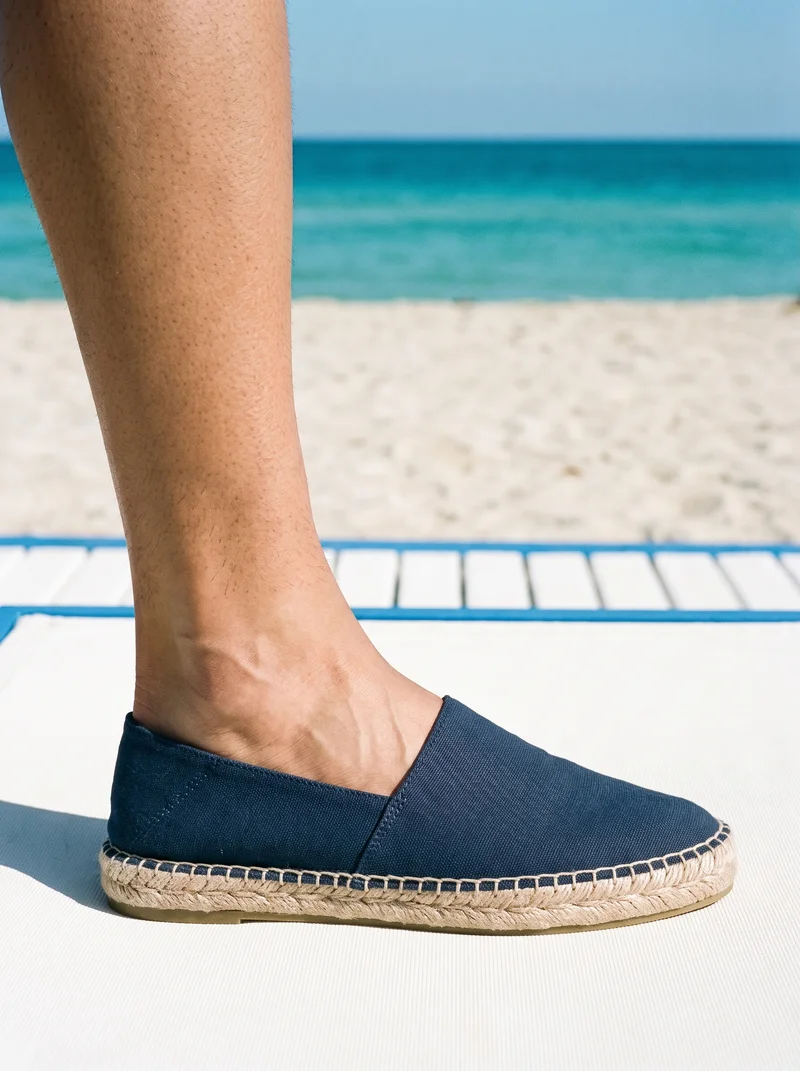 Men's navy canvas espadrilles shown from the side in an ankle-framed view, with the toe pointed. A Greek island beach club serves as the backdrop.