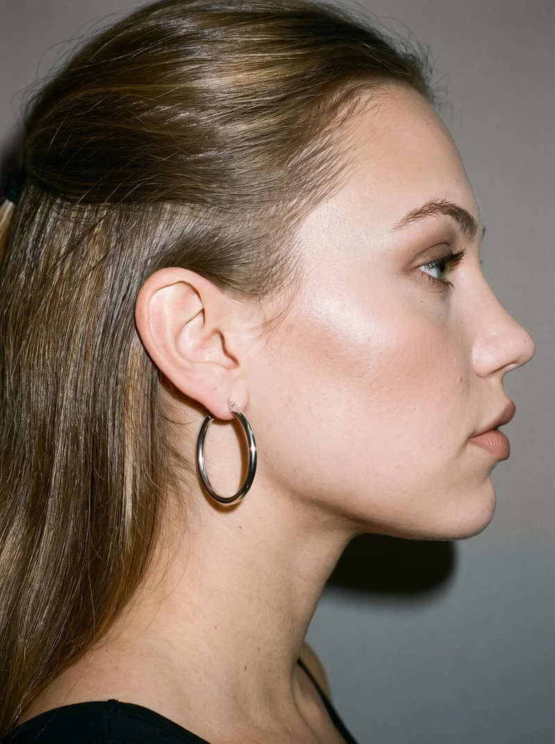 A silver hoop earring worn on an ear, shown in side view with the back of the ear visible. The ear is framed against a studio grey backdrop.