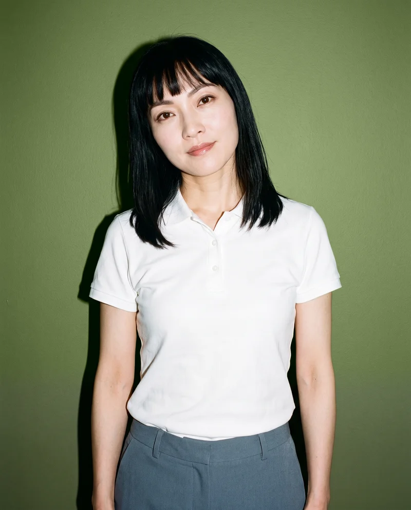 A 37-year-old Chinese woman wears a white polo shirt in an upper-body front view with a shoulder-drop pose and natural expression. An olive grove backdrop is visible behind her.