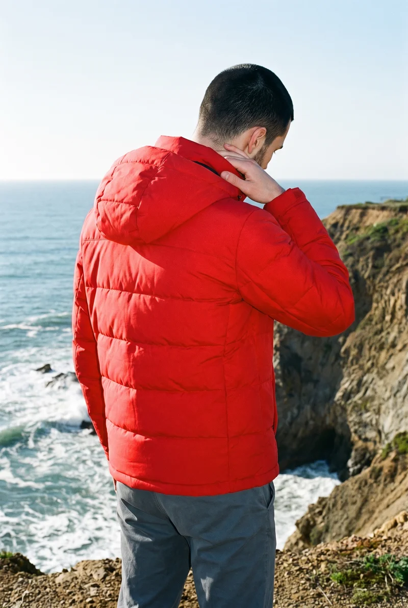 An 18-year-old Caucasian male model is shown from the upper body in a back view, wearing a red insulated ski jacket with one hand at the collarbone. A coastal cliff and Pacific waves form the backdrop.