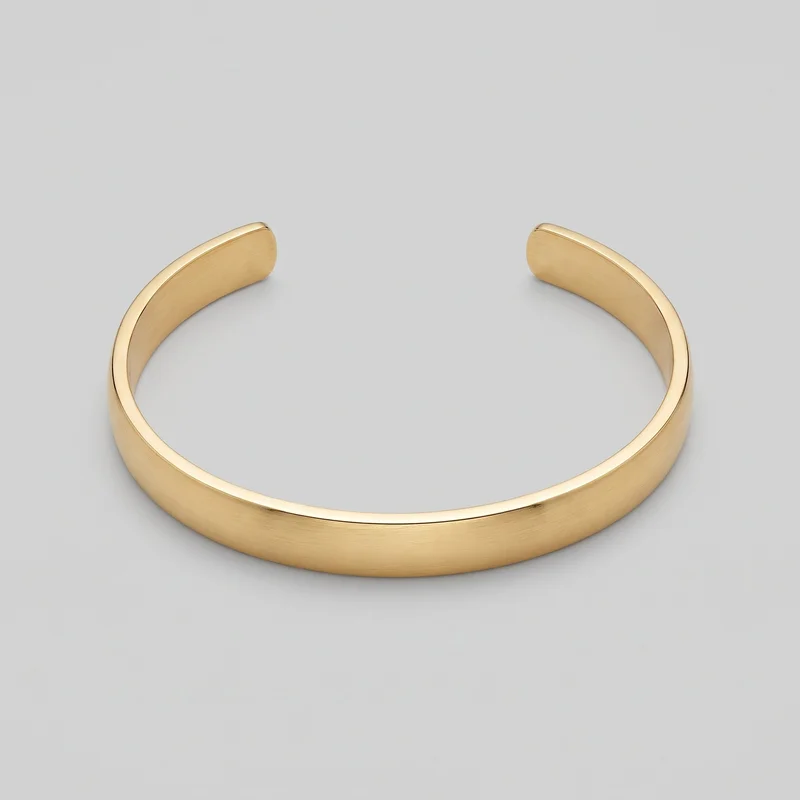 A standalone angled view shows a rigid gold open cuff bracelet with a smooth C-shaped band. The full curved band, open gap, rounded rectangular end caps, raised rounded edges, flatter central surface, and subtle linear texture across the exterior are visible.