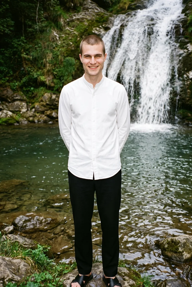 A 22-year-old Caucasian man wears a white grandad shirt in a full-body front view, standing with his hands behind his back and a natural facial expression. A waterfall pool forms the backdrop.