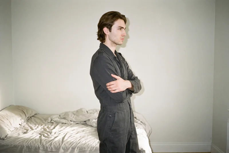 A 22-year-old Caucasian man wears a black boilersuit in a side-view environmental composition. He stands in a self-hug pose with a neutral expression against a bedroom backdrop.