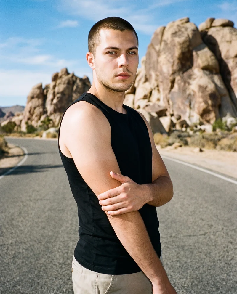 A 21-year-old Caucasian man wears a black vest, shown from the upper body at a front three-quarter angle while holding his elbow with a neutral expression. The Monument Valley Road backdrop is visible behind him.